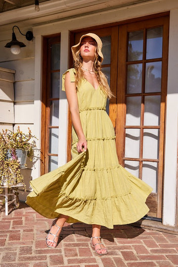 A woman wearing a yellow textured ruffle midi dress with a V-neckline and sleeveless design. She is standing outside, wearing a hat and posing with a confident expression.