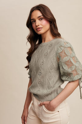 A woman wearing a crochet sleeve pointelle cable knit sweater top in a green color with puff sleeves and a ribbed neckline.