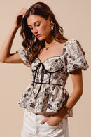Woman wearing floral babydoll corset blouse with puff sleeves, contrast piping, and peplum hem.