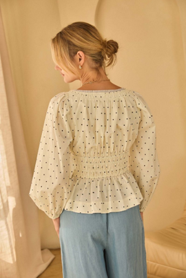 Back view of model wearing a cream polka dot poplin peplum top with puff sleeves and a smocked waist, paired with light blue wide-leg pants.