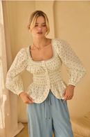 Front view of model wearing a cream polka dot poplin peplum top with square neckline, button front, puff sleeves, and smocked waist, paired with light blue wide-leg pants.
