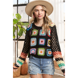 A female model wearing a multicolor crochet sweater with long sleeves, paired with denim shorts and accessorized with a hat and a watch.