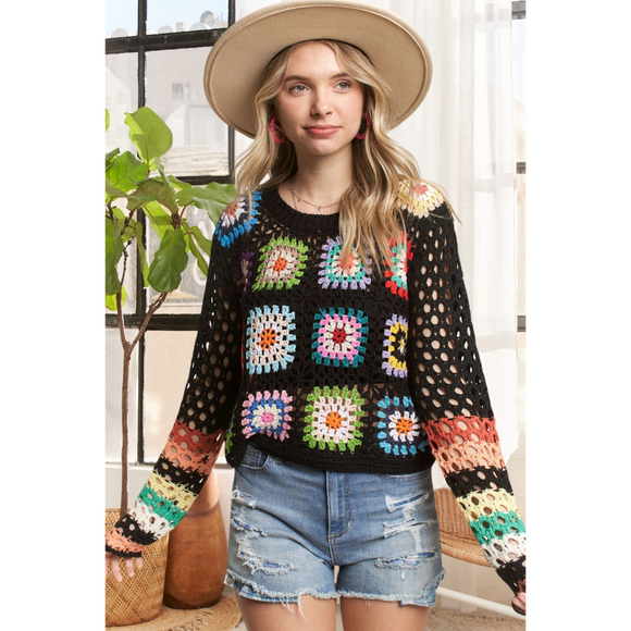 A female model wearing a multicolor crochet sweater with long sleeves, paired with denim shorts and accessorized with a hat and a watch.