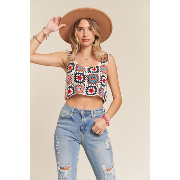 A female model wearing a multicolor crochet crop tank top with spaghetti straps and denim jeans, accessorized with a brown hat and a pink watch.
