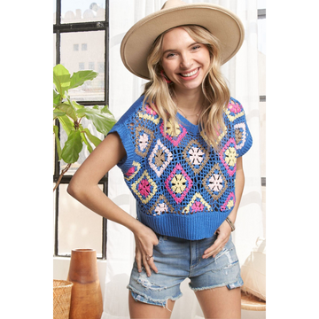 A woman wearing a multi-color crochet vest top with short sleeves, paired with denim shorts and a wide-brimmed hat.