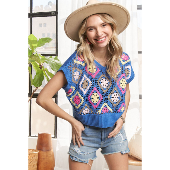 A woman wearing a multi-color crochet vest top with short sleeves, paired with denim shorts and a wide-brimmed hat.