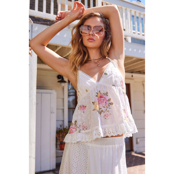 A female model wearing a white sleeveless top with a floral pattern and eyelet details, paired with a white skirt, sunglasses, and a watch.