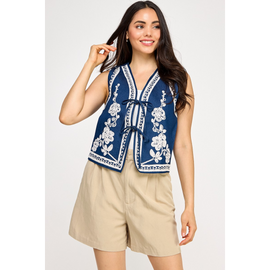 A female model wearing a blue denim vest with white embroidered details and a collar, paired with beige shorts.